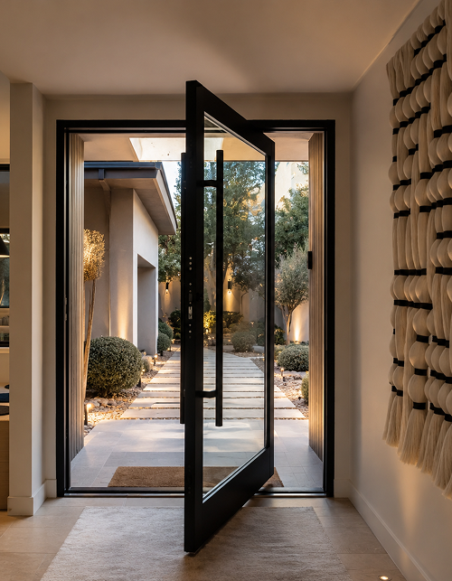 Modern black pivot entry door opening onto a landscaped courtyard walkway